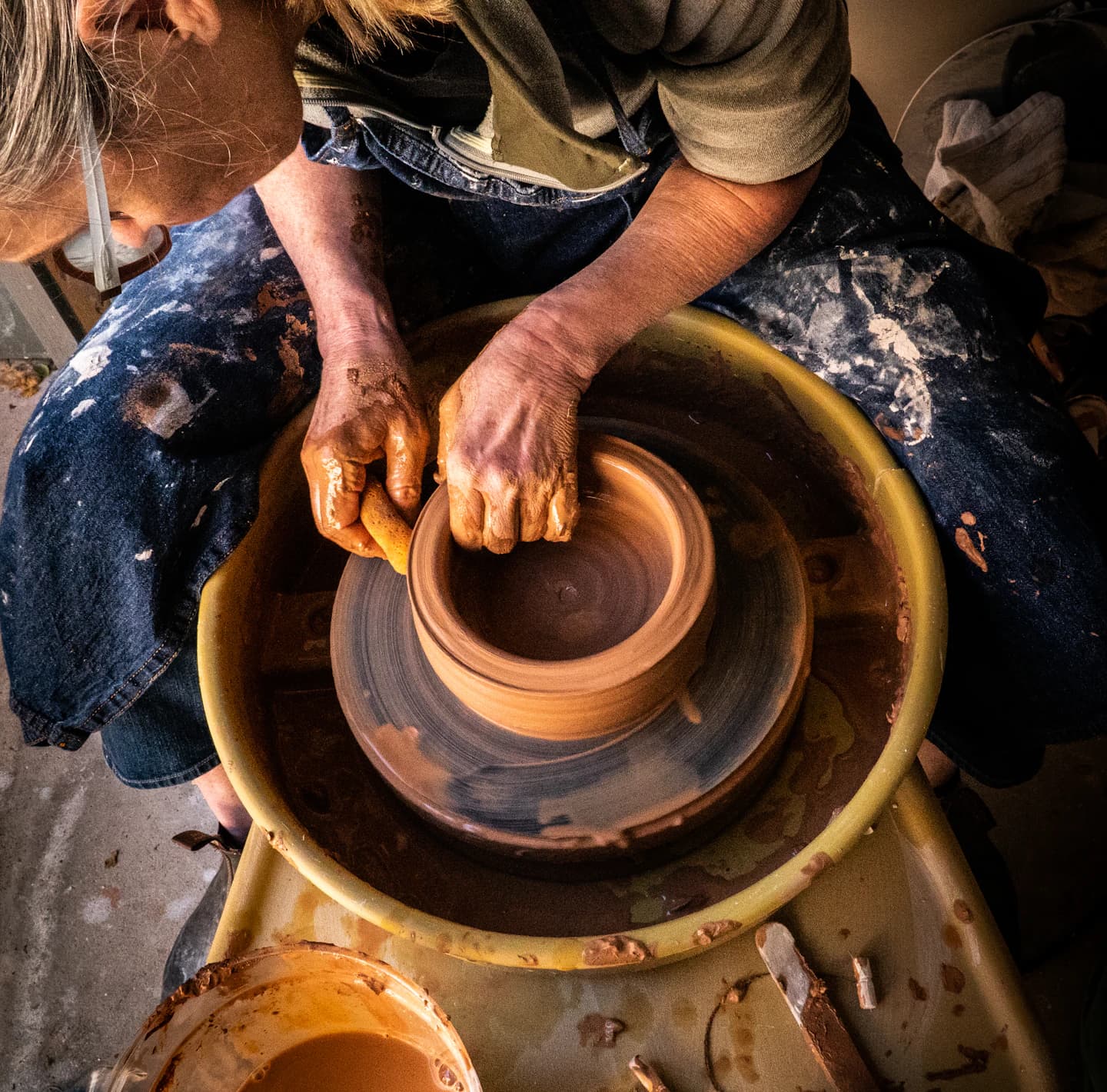 A potter shapes a terracotta bowl on a wheel, wearing clay-marked blue denim.